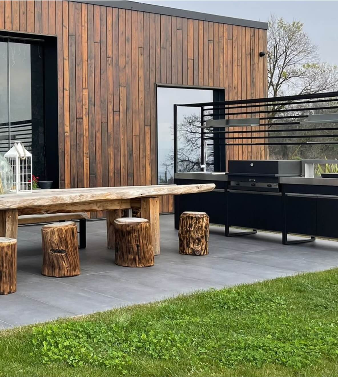 Outdoor kitchen run in black steel with stainless worktop and grill against timber-clad walls, with a live-edge dining table on a paved terrace